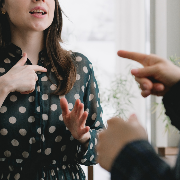 Žena u haljini s točkicama gestikulira dok razgovara, dok druga osoba pokazuje prstom.
