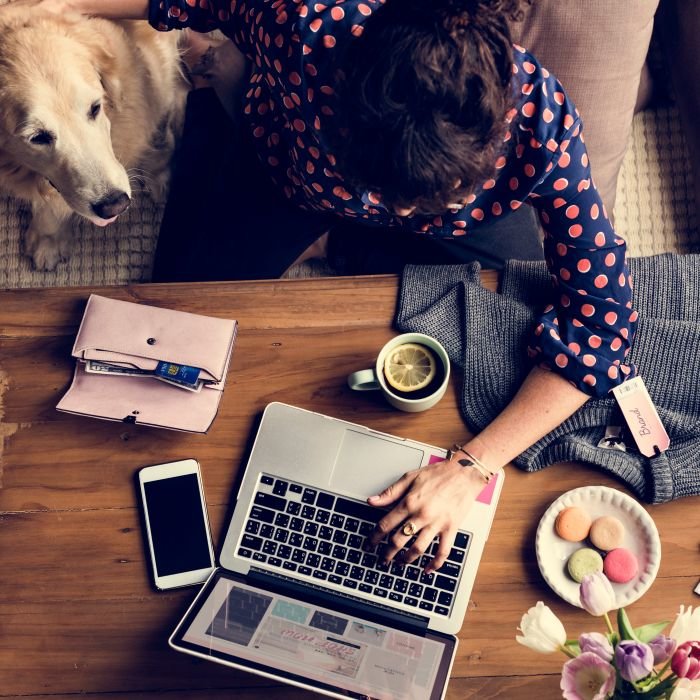 žena s plavom košuljom s crvenim točkicama radi na laptopu, a pored nje je pas kojeg mazi